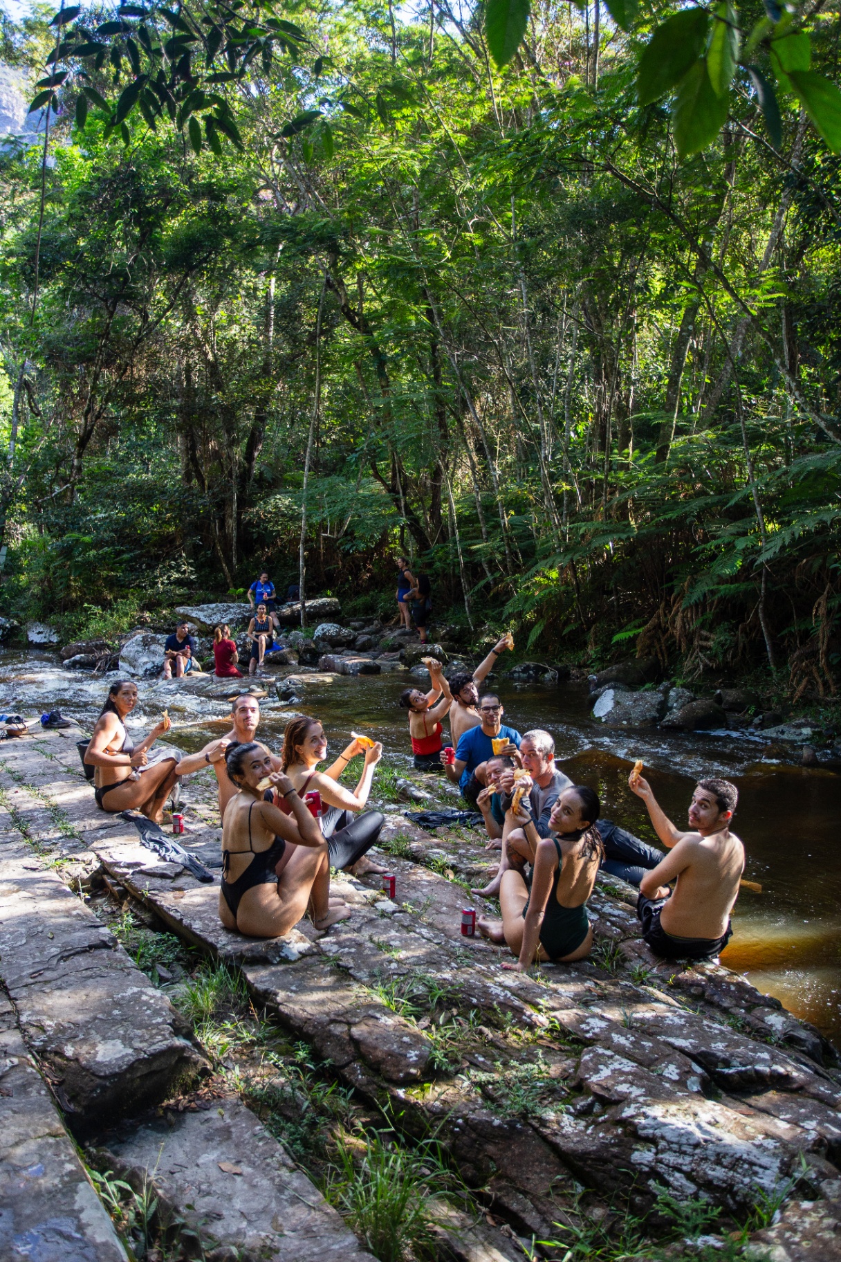 Grupo comendo lanche sentado à beira de um rio na mata