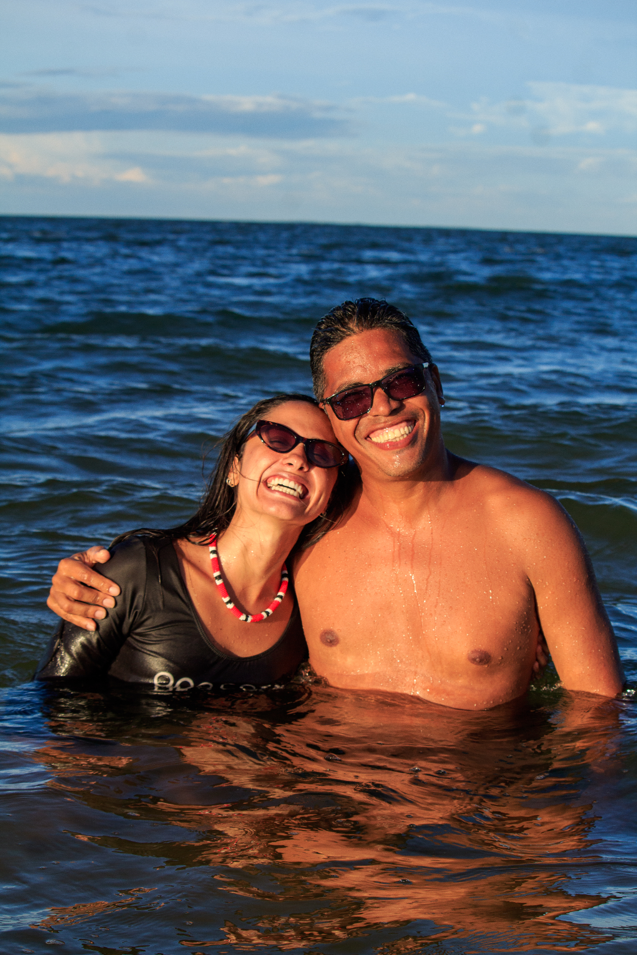 Casal rindo abraçado dentro do mar ao pôr do sol