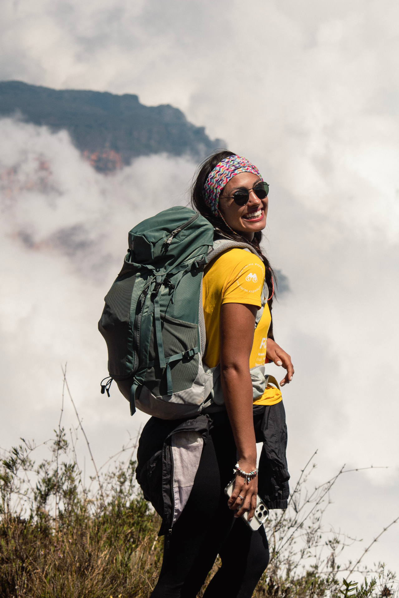 Ana no Monte Roraima com camiseta amarela e mochila de trilha, sorrindo entre as nuvens