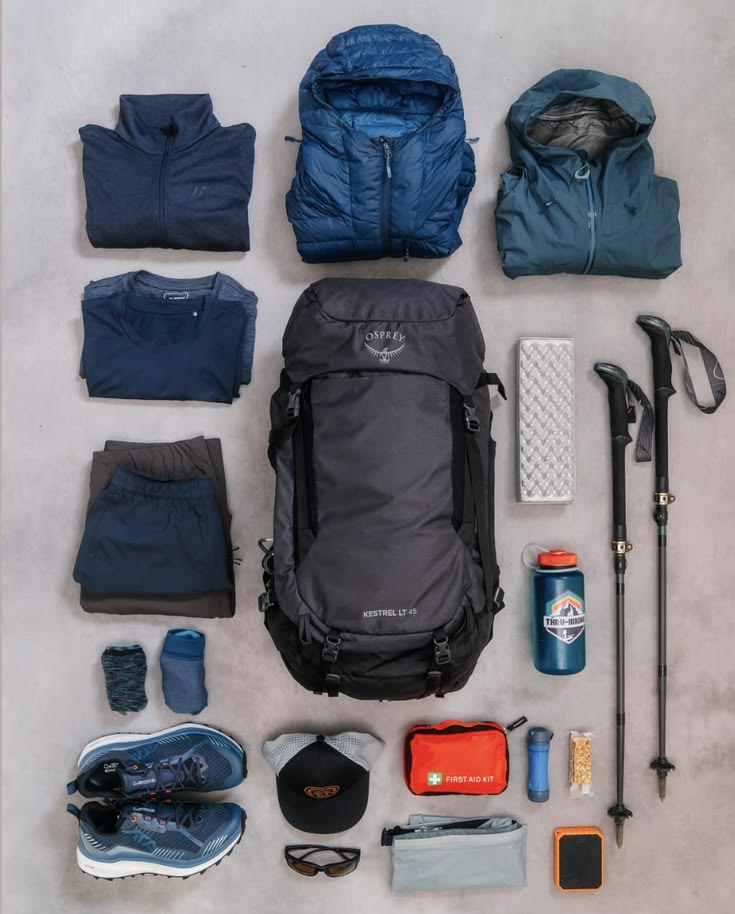 Flat lay do kit completo pra expedição: mochila Osprey, bastões, kit primeiros socorros, tênis, garrafa, boné e roupas técnicas em tons de azul