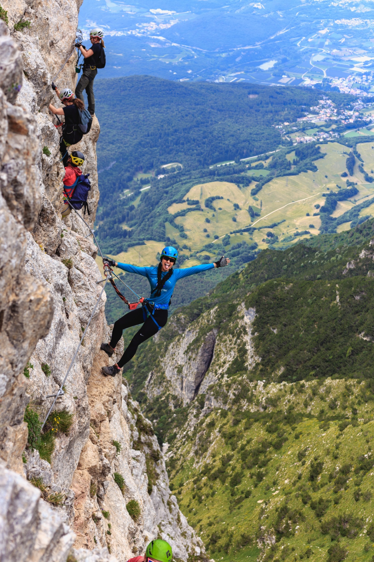 Ana pendurada rindo numa via ferrata nos Alpes, com o vale verde lá embaixo